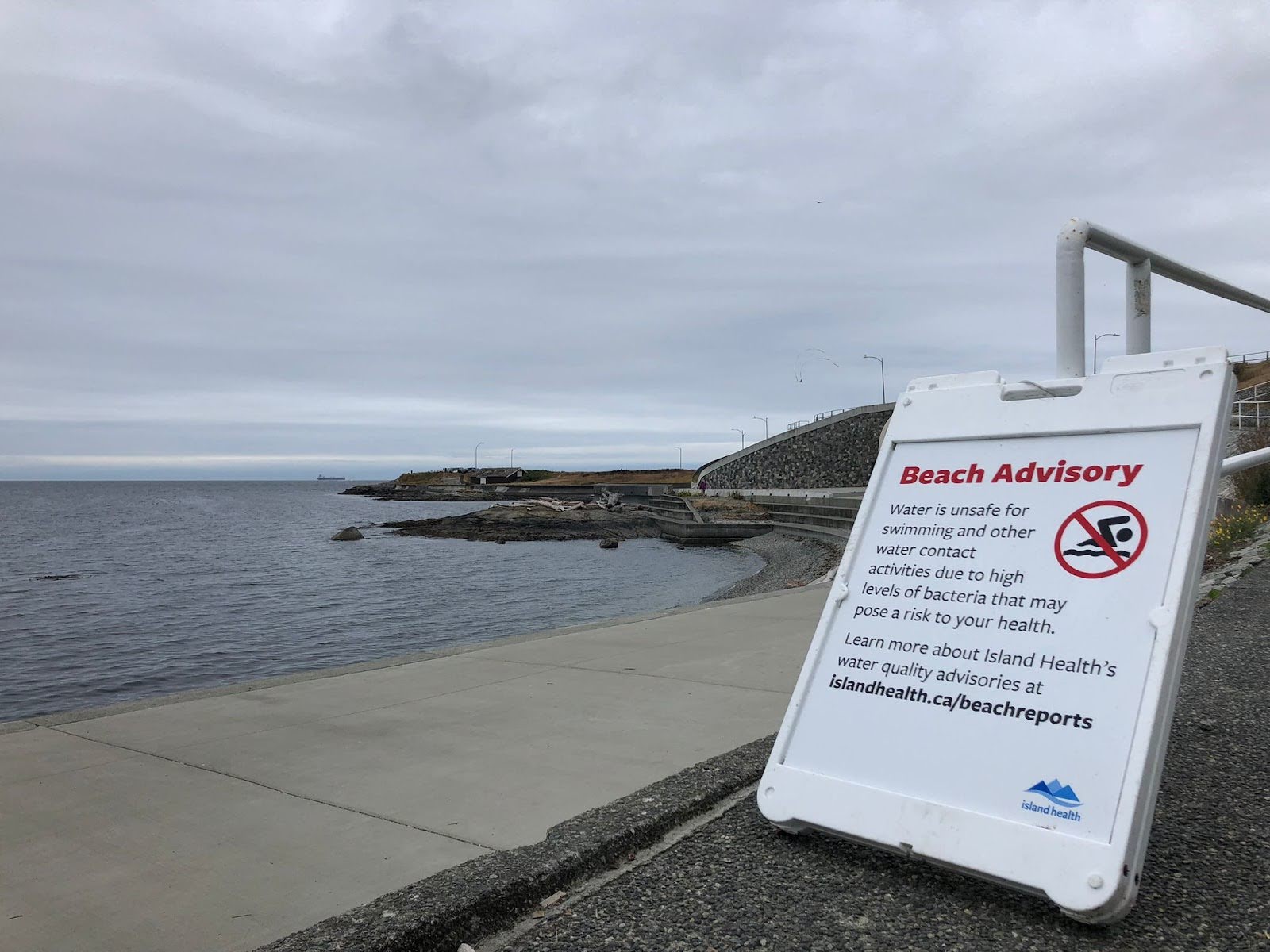 A beach advisory warning people that water is unsafe for swimming and other water contact at Ross Bay Beach. The Clover Point Pump Station is in the background. 